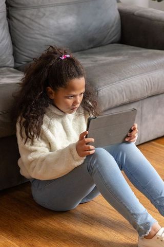 Concentrating school-age girl sitting on floor using tablet, cozy fleece, home learning