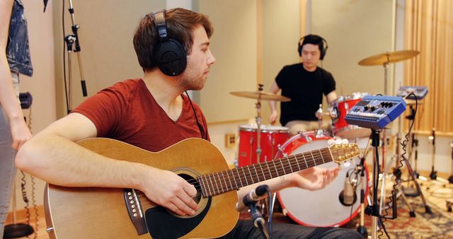 Musicians rehearsing in recording studio. One is playing an acoustic guitar while another plays the drums. Both are wearing headphones and seem focused on their work. Ideal for use in content related to music production, band rehearsals, recording sessions, and creative collaborations.