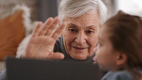 Senior woman waving to laptop while grandchild making funny face on living room sofa