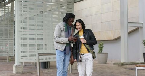Multicultural friends walking plaza checking smartphone holding coffee and skateboard