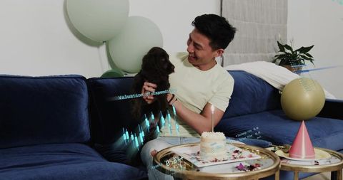 Young man holding puppy celebrating birthday on sofa with cake, balloons and holographic charts