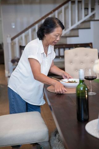 Senior asian woman placing bowl on dining table with wine, preparing casual family dinner