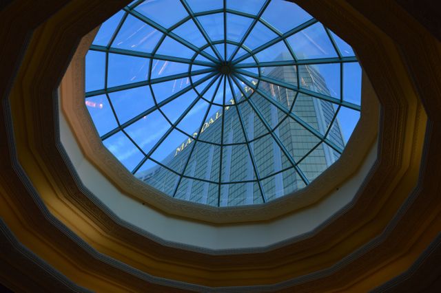 Photo showcasing a modern skyscraper viewed through an intricate glass roof from the interior of a building. The geometric design of the roof frames the towering structure, creating a visually striking composition. Ideal for use in content related to architecture, structural engineering, urban development, modern living, or real estate promotion.