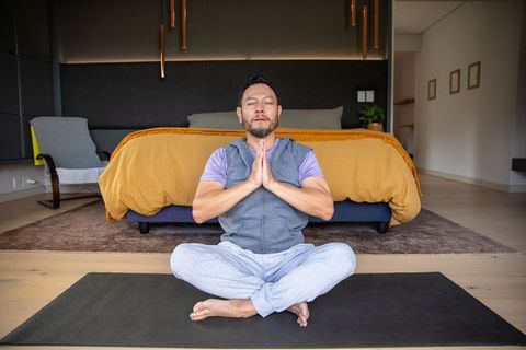 Mature asian man meditating barefoot on yoga mat in contemporary bedroom with mustard bedding