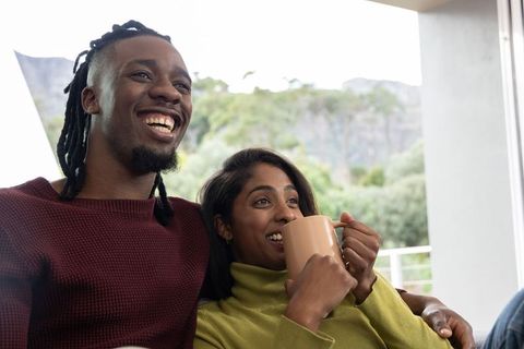 Laughing diverse couple cuddling on sofa holding beige mug enjoying cozy balcony morning
