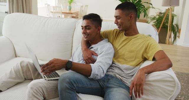 Smiling biracial gay male couple sitting on sofa looking at laptop and talking. staying at home in isolation during quarantine lockdown.