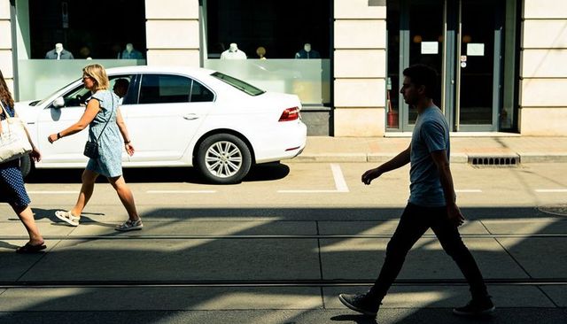 Man walking across sunlit city street past white sedan and storefront in long shadows