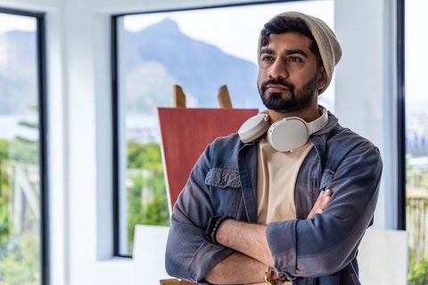 Thoughtful indian artist wearing beanie and headphones standing by easel in modern studio