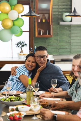 Multicultural friends hugging at festive home dinner with wine, balloons, laughter