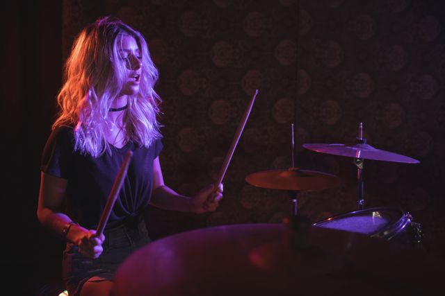 Confident female drummer performing on an illuminated stage in a nightclub. Ideal for use in articles or promotions related to live music, nightlife, female musicians, and entertainment events. Can also be used in advertisements for music venues, concerts, and musical instruments.