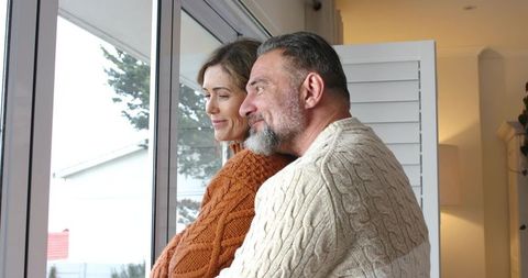 Middle-aged couple embracing by window, wearing cozy cable-knit sweaters, gazing outside