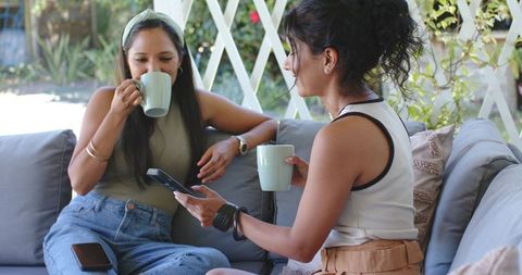 Indian and African American friends sharing coffee on patio sofa, relaxing with phone