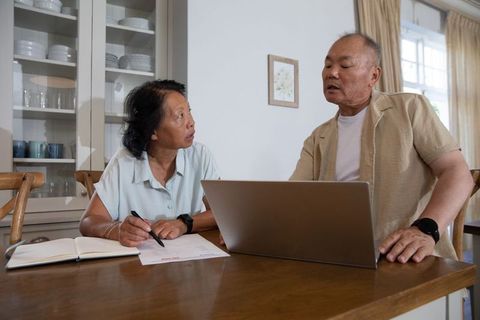 Senior asian couple discussing finances at kitchen table using laptop and paperwork