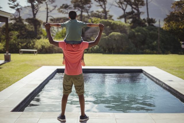 Father carrying son on shoulders near poolside in backyard. Ideal for depicting family bonding, summer activities, outdoor fun, and parenting moments. Suitable for advertisements, family-oriented content, and lifestyle blogs.