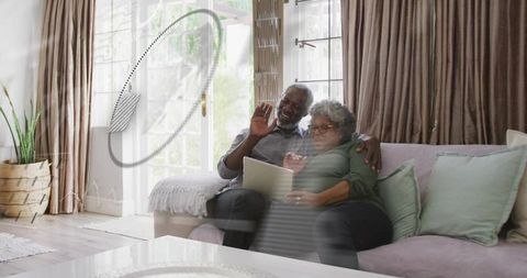 Senior couple waving on tablet video call while cuddling on cozy sunlit living room sofa