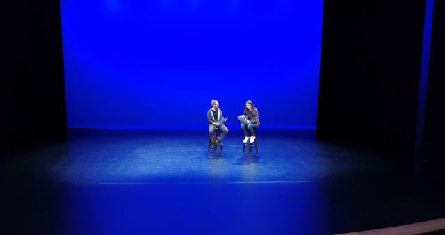 A Caucasian man and woman sit on stage under a blue spotlight, with copy space. Their relaxed posture suggests a casual conversation or rehearsal break in a theater setting.