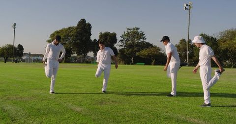 Four cricket players stretching and balancing on green field during pre-match warmup