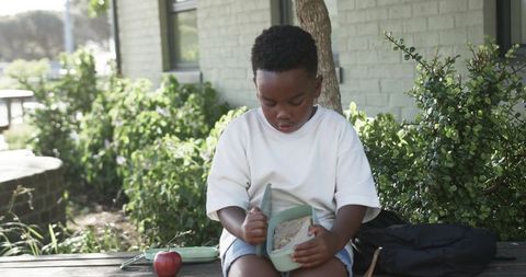 African american boy sitting outdoors eating lunch from green container in schoolyard