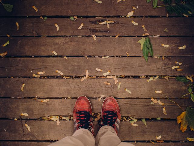 Top view of person standing in red shoes on wooden deck scattered with leaves. Ideal for use in autumn-themed projects, lifestyle blogs, or nature-related content. Suitable for conveying a casual and laid-back atmosphere.