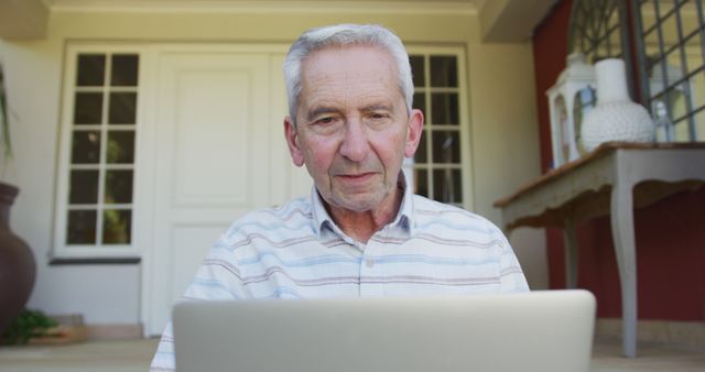 Senior man sitting on porch using laptop. Useful for depicting seniors embracing technology, internet usage among elderly, active retirement lifestyle, or home office setups.