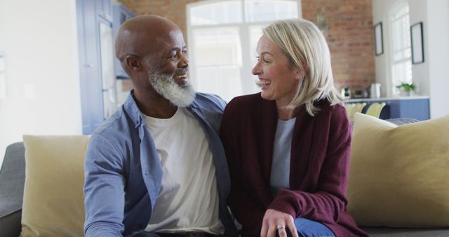 This heartwarming photo captures a happy multiracial couple sharing a laugh while sitting on a cozy sofa at home. The relaxed and intimate atmosphere makes this image perfect for illustrating themes of love, togetherness, relationships, and lifestyle in various contexts, such as promotional materials, social media posts, web articles, and blogs about relationships, home life, and happiness.