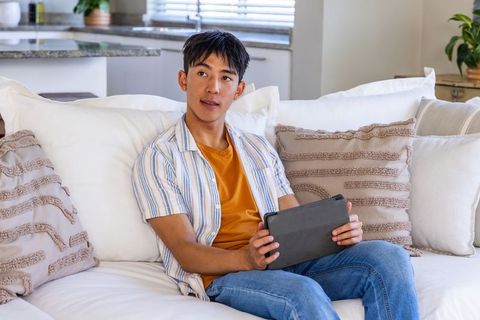 Asian man relaxing on white sofa holding tablet in sunlit modern living room with cushions