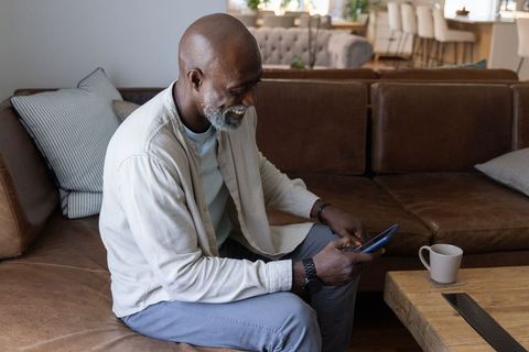 Senior african man relaxing on sofa using smartphone with coffee in modern living room
