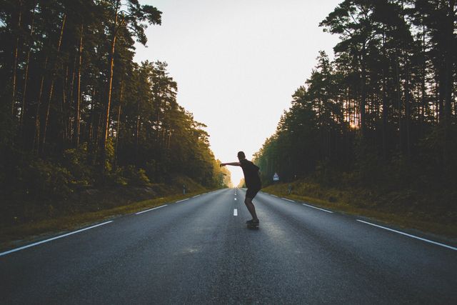 Person skateboarding on an empty, tree-lined road during sunset, surrounded by nature. Perfect for use in travel blogs, adventure advertising, promoting outdoor activities, or lifestyle imagery illustrating solitude, exploration, and the freedom of road trips.