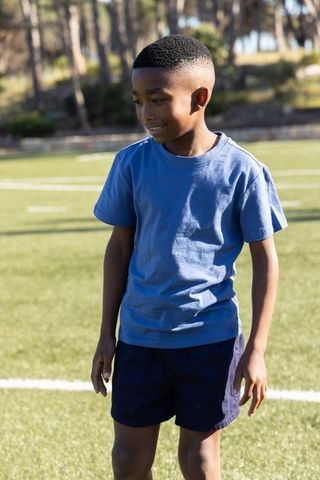 African american boy standing on turf wearing blue t-shirt navy shorts sunny sports field