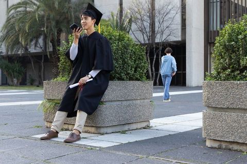 Asian graduate sitting on campus planter talking on mobile holding diploma in cap and gown
