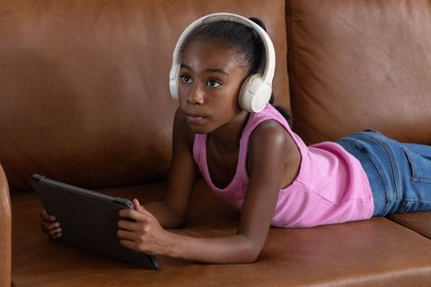 Young girl lying on couch wearing white headphones using tablet for study and leisure
