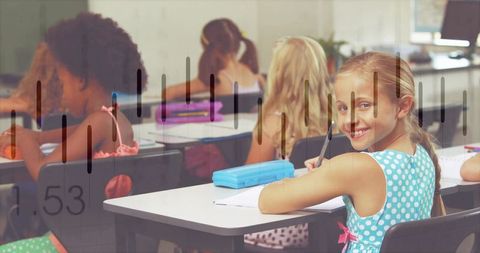 Smiling blonde girl writing at school desk in turquoise polka dot dress with classmates