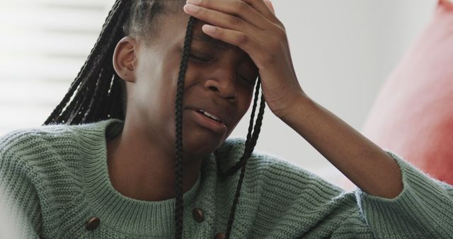 Young girl in green sweater showing distress, touching her forehead while crying. Useful for materials focused on mental health, stress, trauma, and emotional well-being.