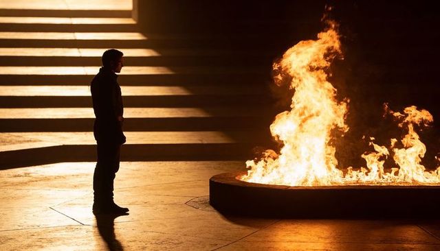 Silhouetted man standing facing glowing oval fire pit on plaza steps at night, amber light