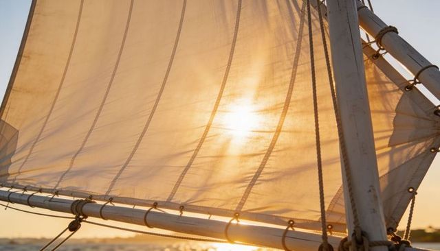 Backlit mainsail catching golden sunset light on small sailboat boom and rigging, translucent canvas