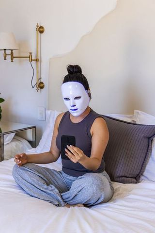 Woman wearing led facial mask on bed using smartphone for at-home skincare routine
