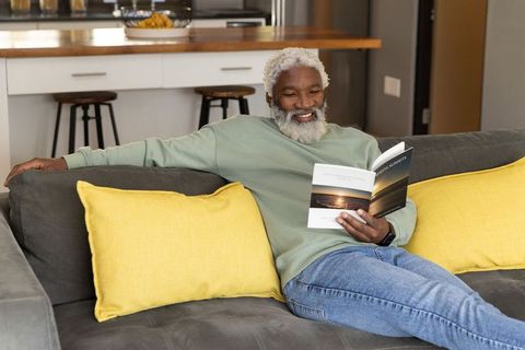 Senior african american man reclining on dark gray sofa reading paperback, cozy home relaxation