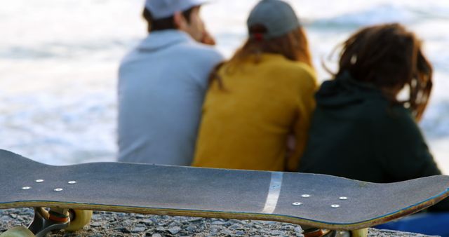 Three friends sitting and relaxing near the ocean, with a skateboard prominently placed in the foreground. This image captures relaxed outdoor moments and youthful lifestyles. It can be used for social media, lifestyle blogs, or marketing content related to outdoor activities and friendship.