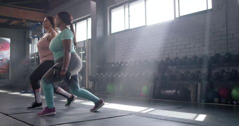 Two women performing dumbbell lunges in sunlit industrial gym for strength training