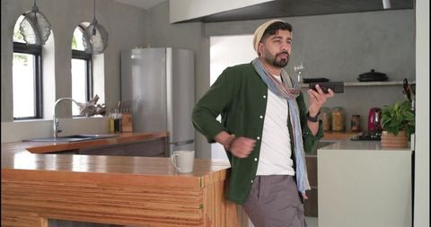 Indian man leaning on modern rustic kitchen island using smartphone enjoying morning coffee