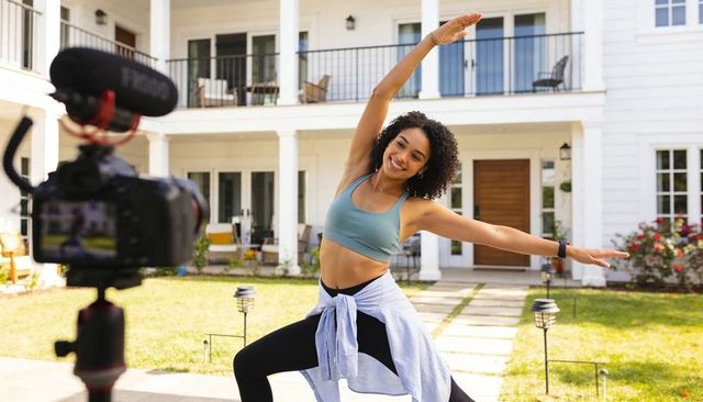 Fitness creator filming outdoor yoga lunge on front lawn with camera and microphone