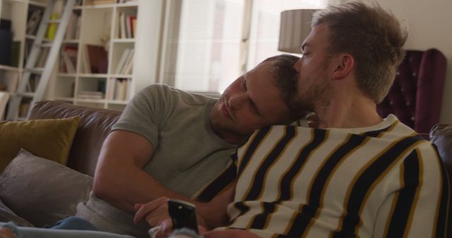 Happy caucasian gay male couple sitting on couch, embracing and looking tv in living room. Togetherness, relationship, romance and domestic life, unaltered.