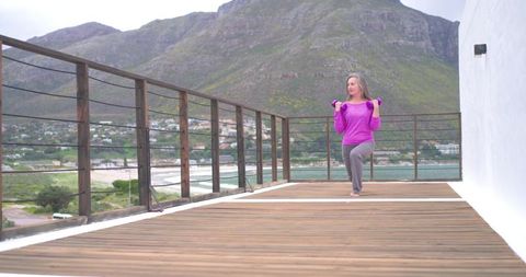 Mature woman exercising with resistance band on seaside wooden terrace with mountain view