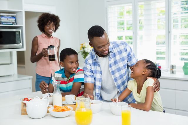 African American family enjoying breakfast together in a bright kitchen. Parents and children are smiling and interacting, creating a warm and joyful atmosphere. Ideal for use in advertisements, family-oriented content, lifestyle blogs, and parenting articles.