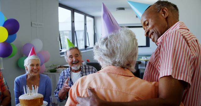 Group of elderly friends celebrating a birthday at a festive gathering. They are wearing party hats, surrounded by balloons, and a birthday cake with candles is in front of them. This image can be used in contexts related to elderly celebrations, senior communities, birthdays, friendship, and joyful gatherings.