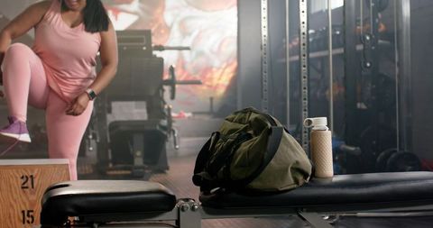 African american woman performing plyo step-up in pink activewear with duffel bag on bench