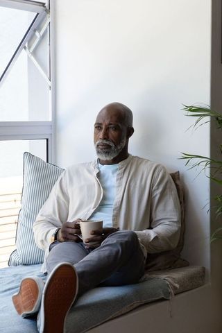 Senior african man sitting on window bench holding coffee mug, calm minimalist portrait