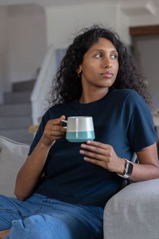Indian woman relaxing on sofa holding two-tone mug and wearing smartwatch in cozy interior