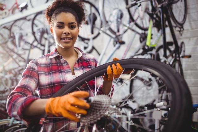 Female mechanic holding and repairing a bicycle wheel in a workshop. She is wearing orange gloves and a plaid shirt, surrounded by various bicycles and tools. Ideal for use in articles or advertisements related to bicycle repair services, women in trades, or mechanical workshops.