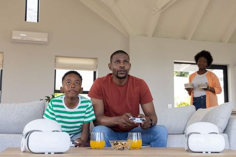 African american father and son playing video games on sofa with vr headsets